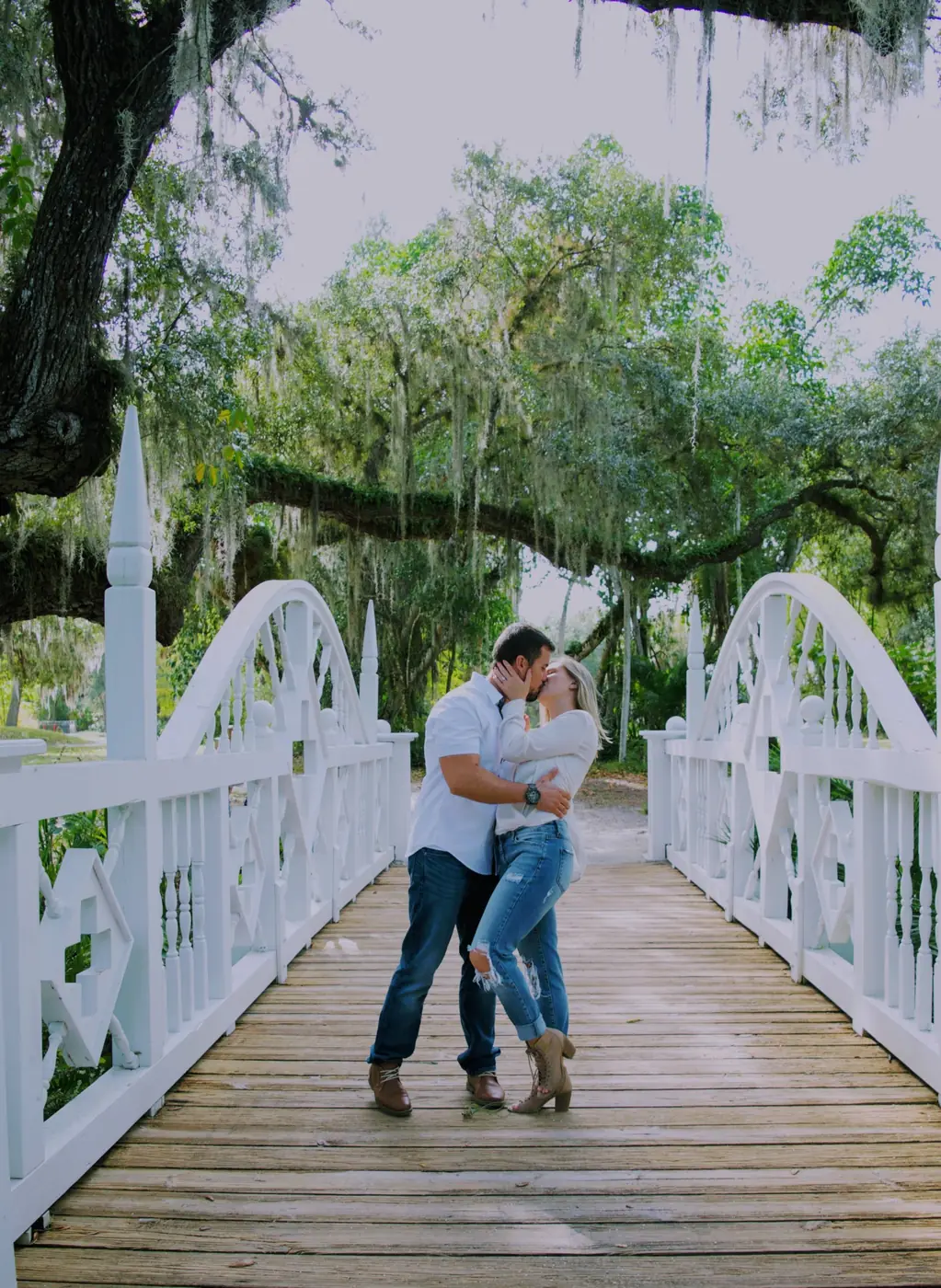 Engaged couple kissing on a white bridge under oak trees