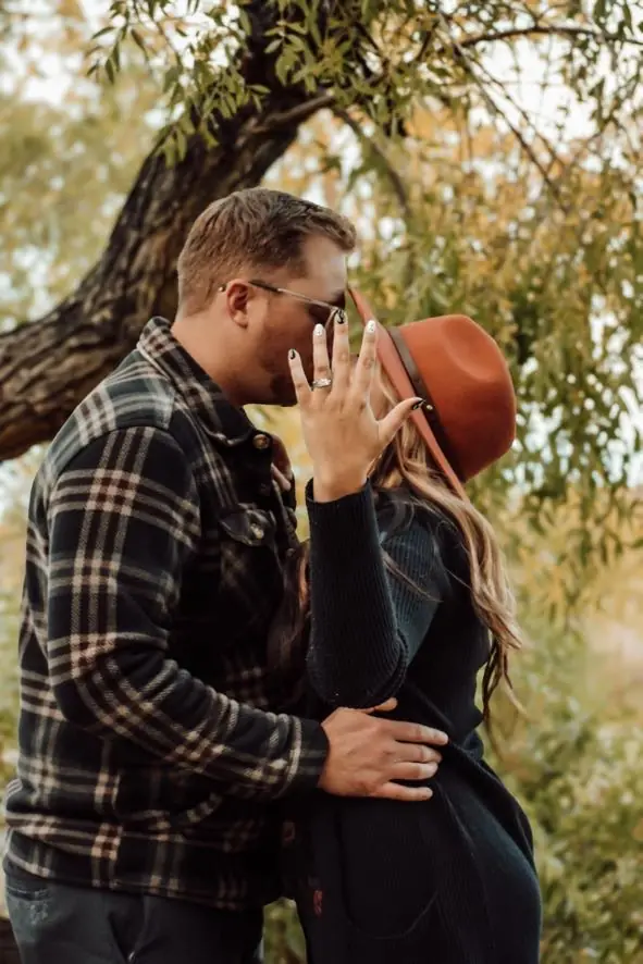 Engaged couple kissing under a willow tree as she shows her ring