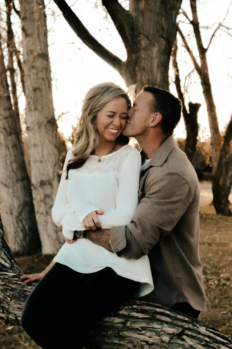 Engaged couple on a tree trunk at sunset, she laughs as he kisses her cheek