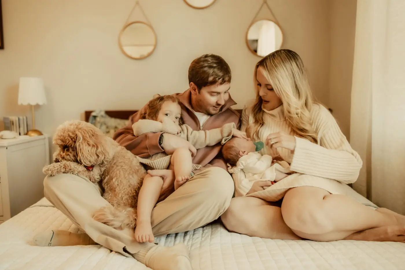 Parents cuddling their toddler and newborn on the bed with the family dog