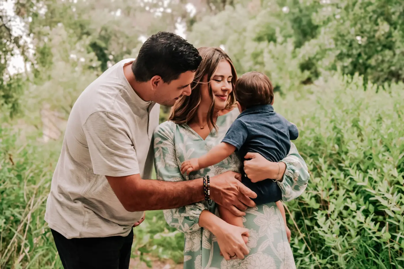 Parents leaning in to hug their baby in a leafy green path