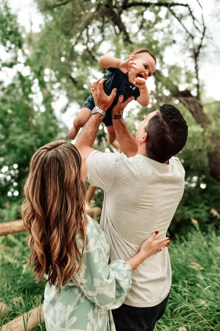 Dad lifting his laughing baby into the air with mom beside him in the trees