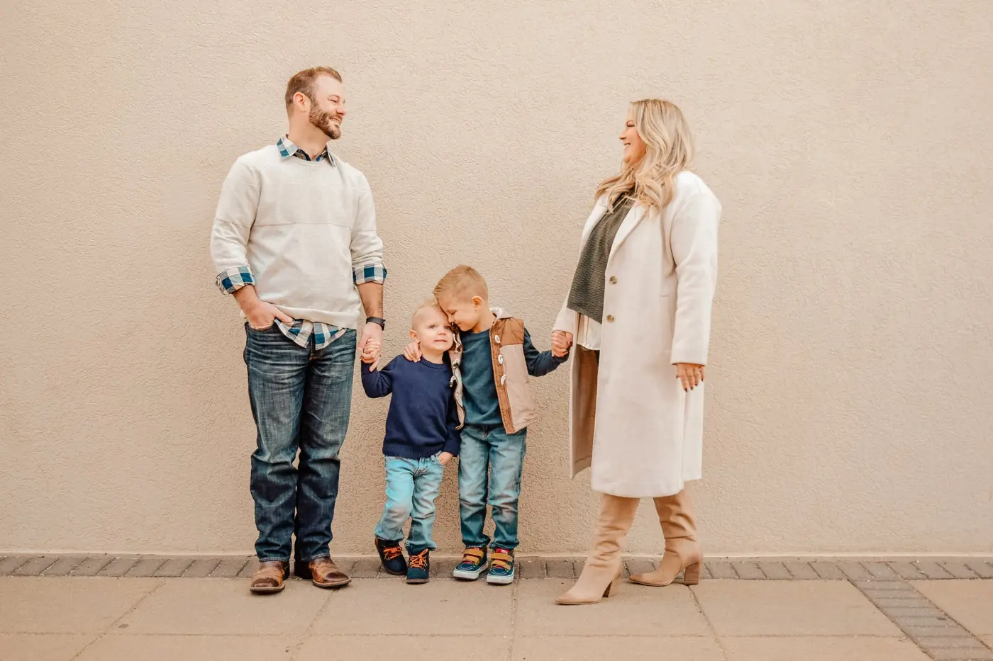 Parents holding hands with their two little boys in front of a cream wall