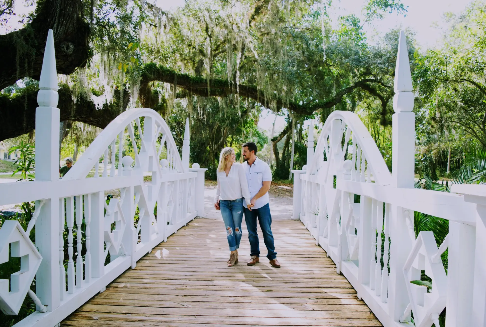 Engaged couple walking hand in hand across a white bridge beneath moss-draped oak trees