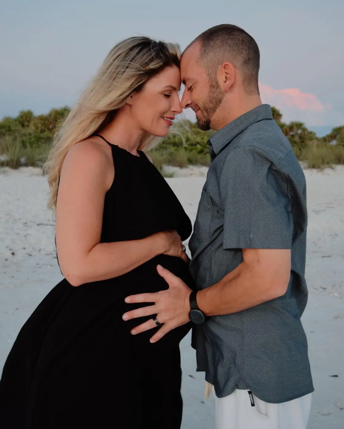 Expecting couple touching foreheads on the beach at dusk, hands on her belly