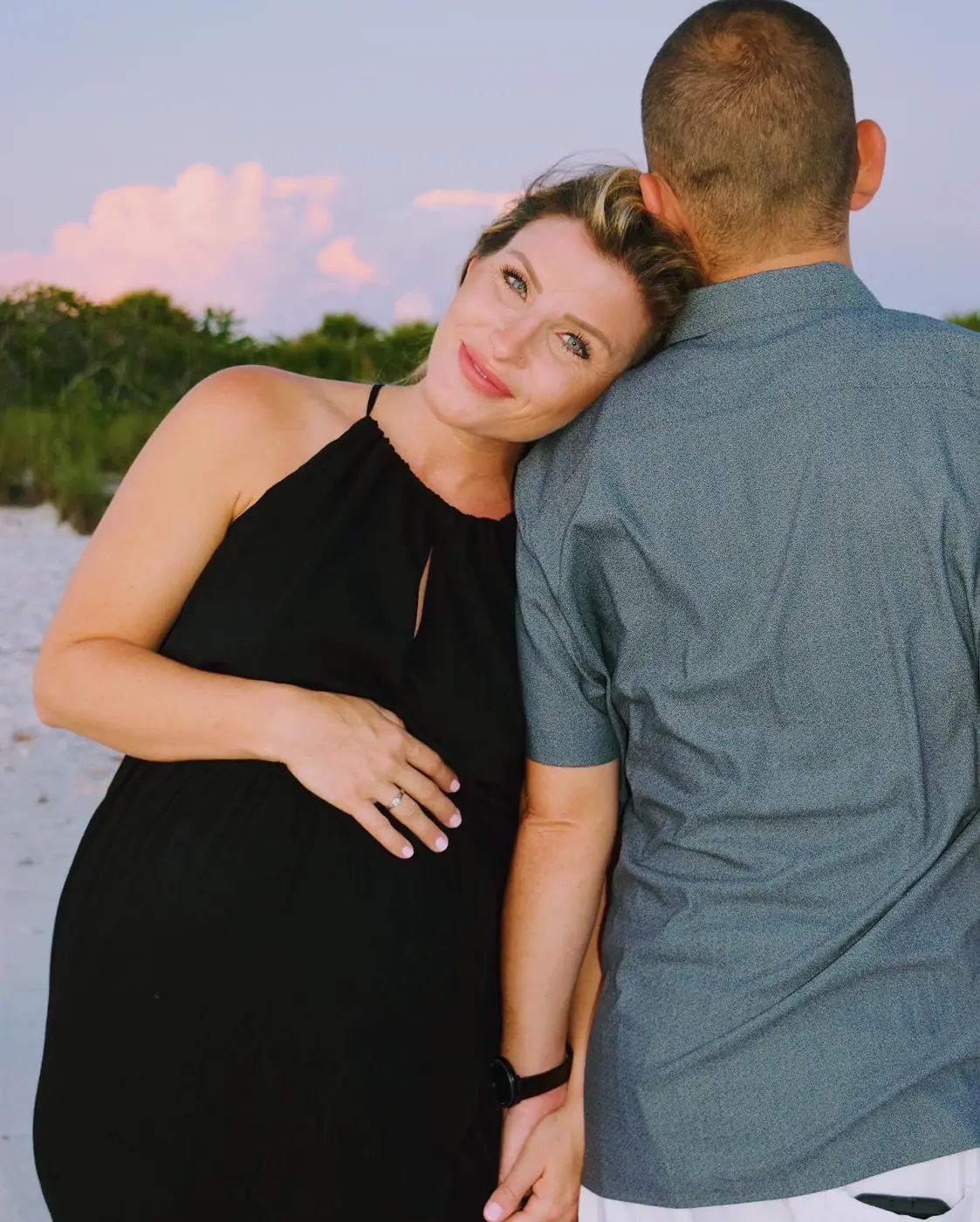 Expecting mother in black leaning her head on her husband’s shoulder at the beach