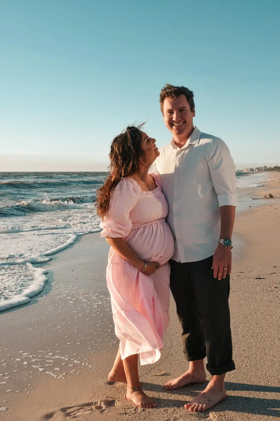 Expecting mother in a pink dress laughing up at her husband on the beach