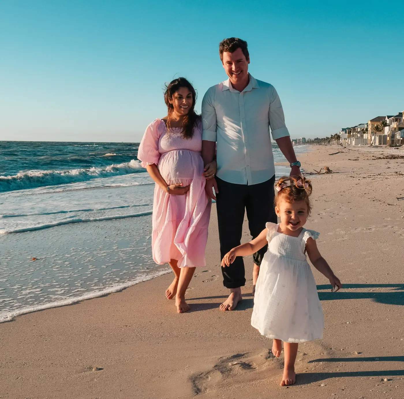 Expecting parents walking the beach in the sunset as their toddler runs ahead
