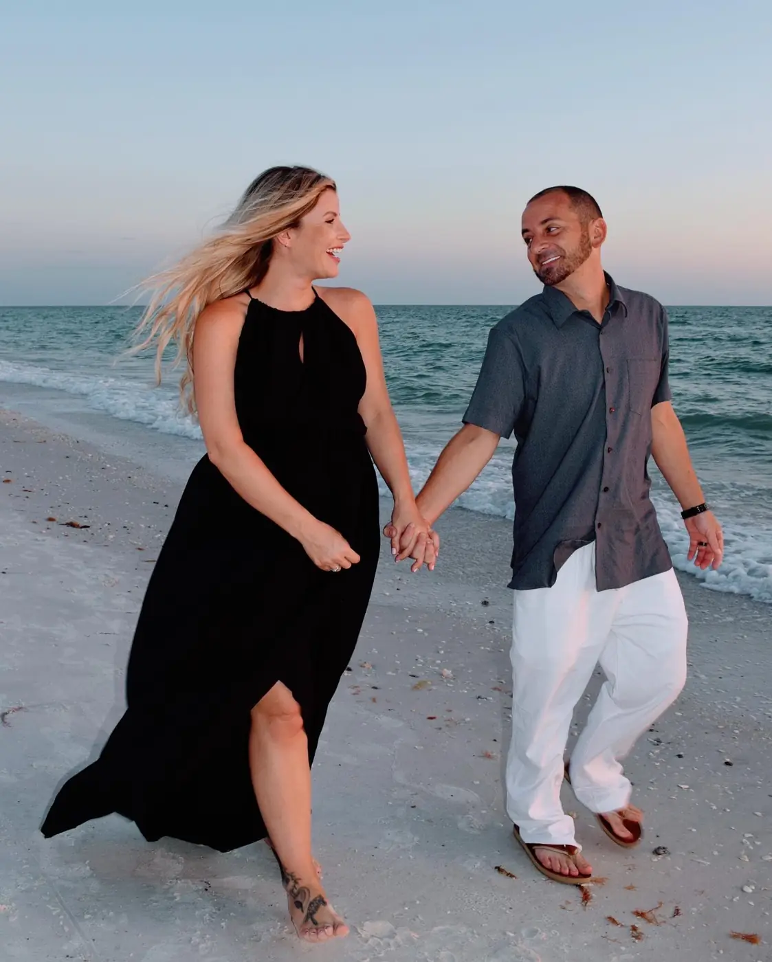 Expecting couple holding hands and laughing as they walk along the shoreline