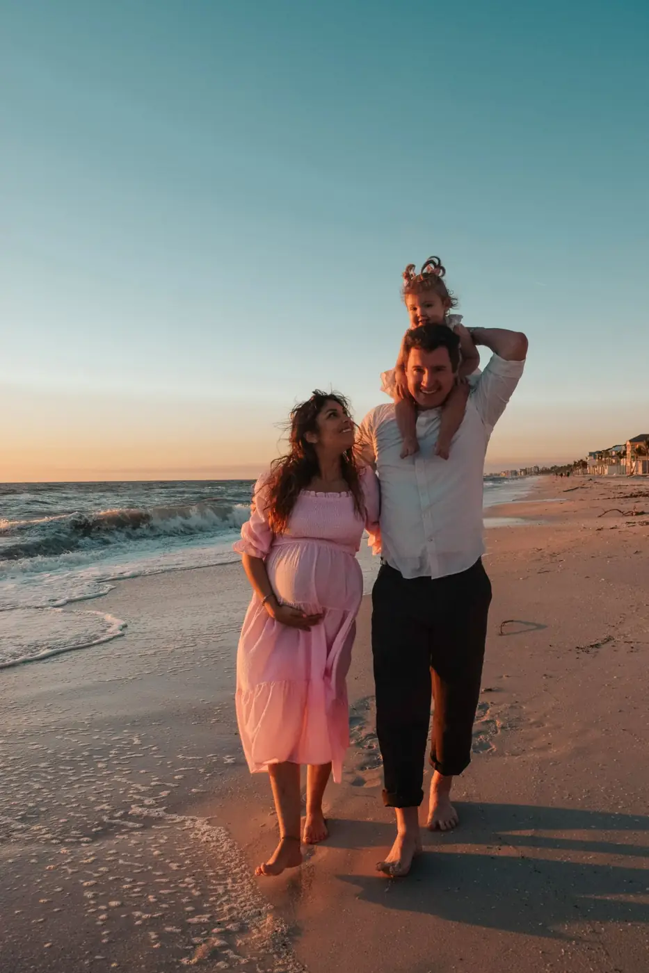 Dad carrying his toddler on his shoulders as his expecting wife looks up at them on the beach
