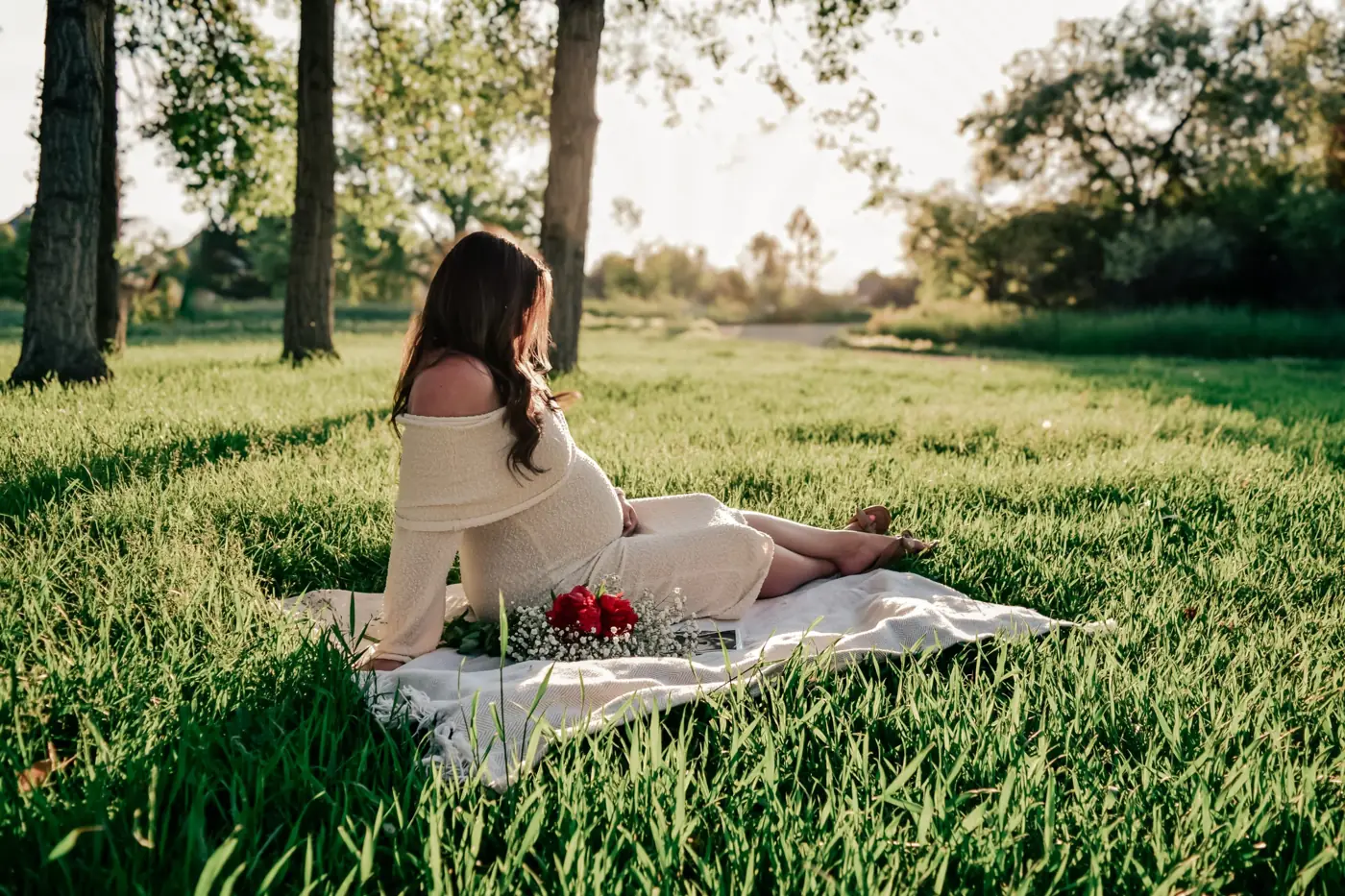 Expecting mother in a cream dress sitting on a blanket with red roses, looking out over the grass