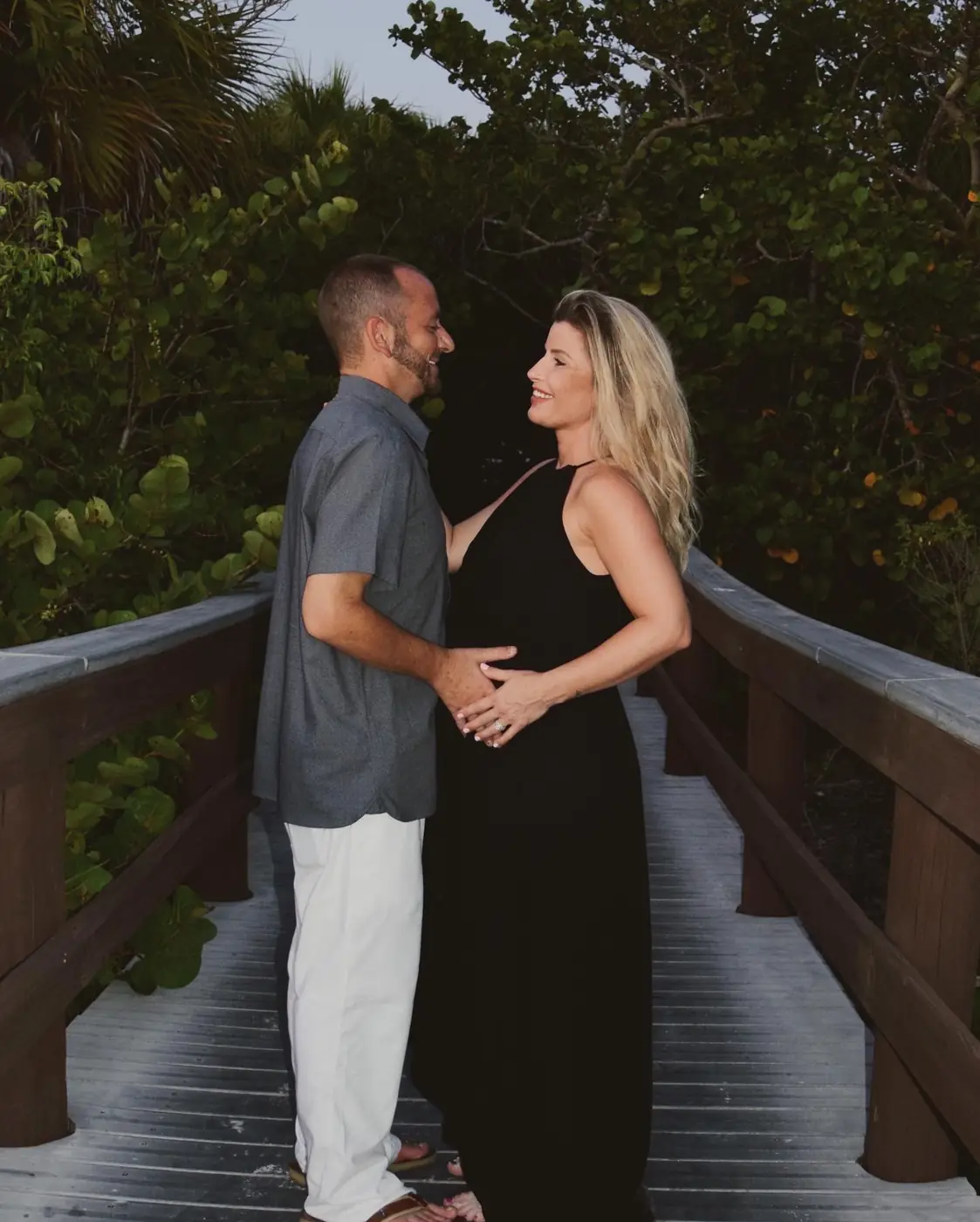 Expecting couple smiling at each other with hands on her belly on a beach boardwalk