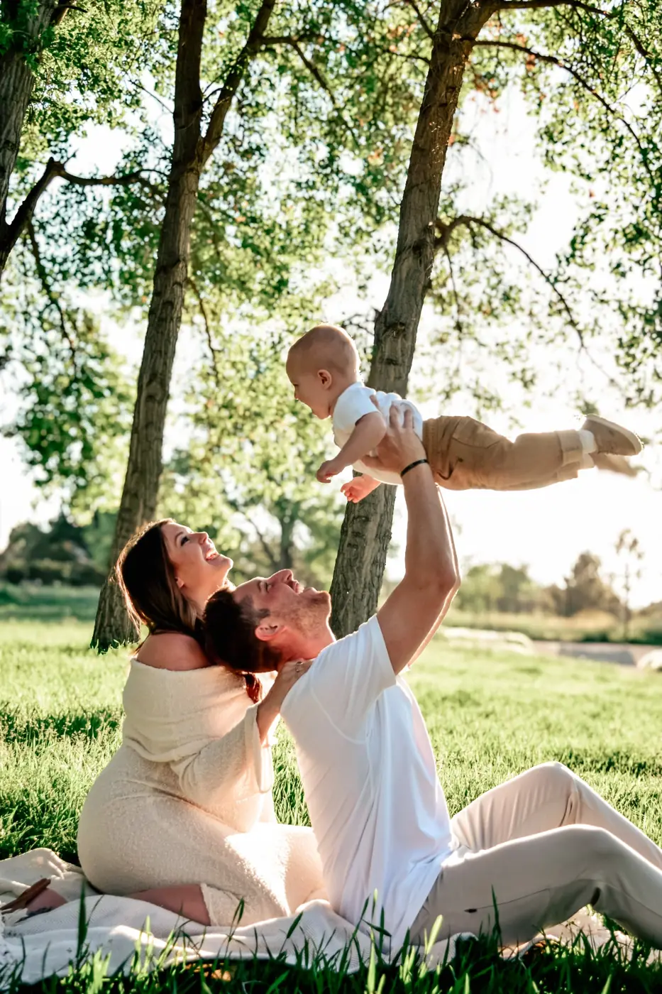 Dad lifting his toddler overhead as his expecting wife laughs on a blanket under the trees