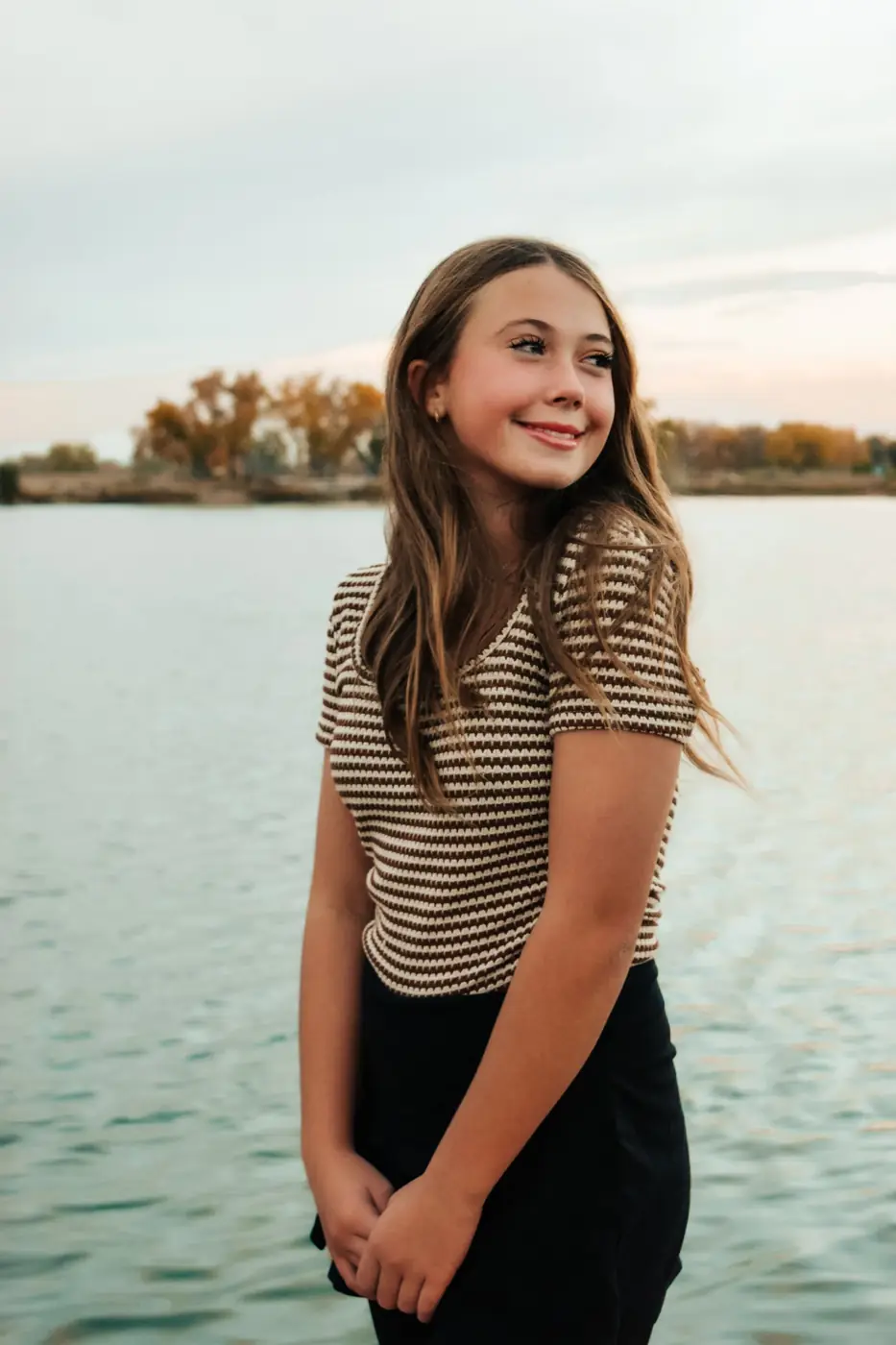 Senior girl in a striped top smiling over her shoulder by the lake at sunset