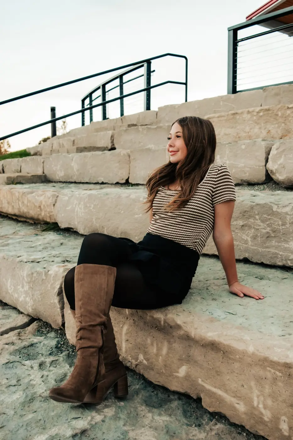 Senior girl in a striped top and tall boots sitting on stone steps, looking off to the side